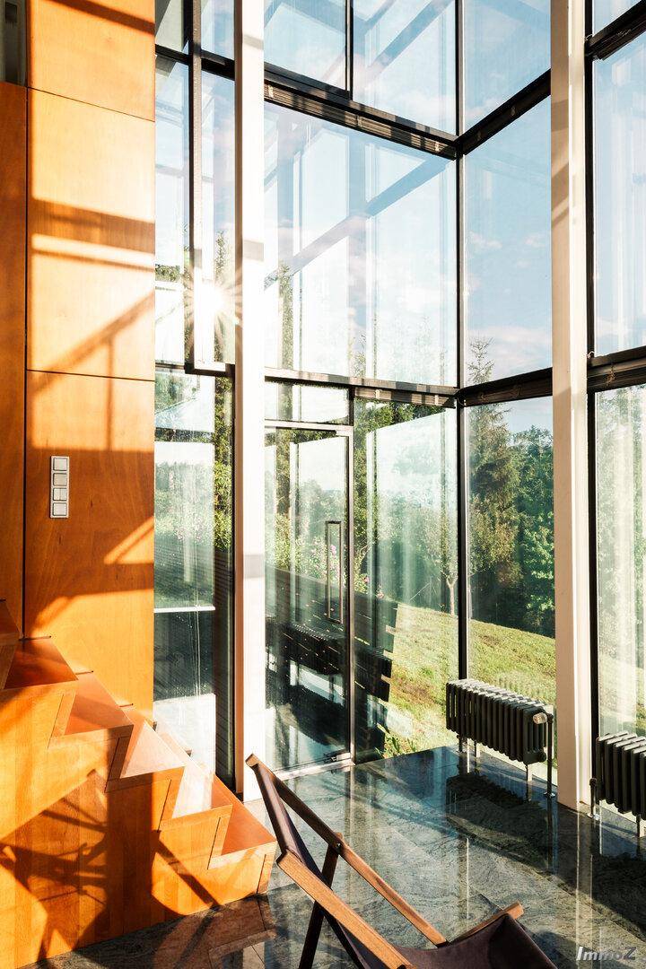 Lichtdurchfluteter Bereich mit hoher Glasfassade, Holztreppe und Blick ins Grüne.