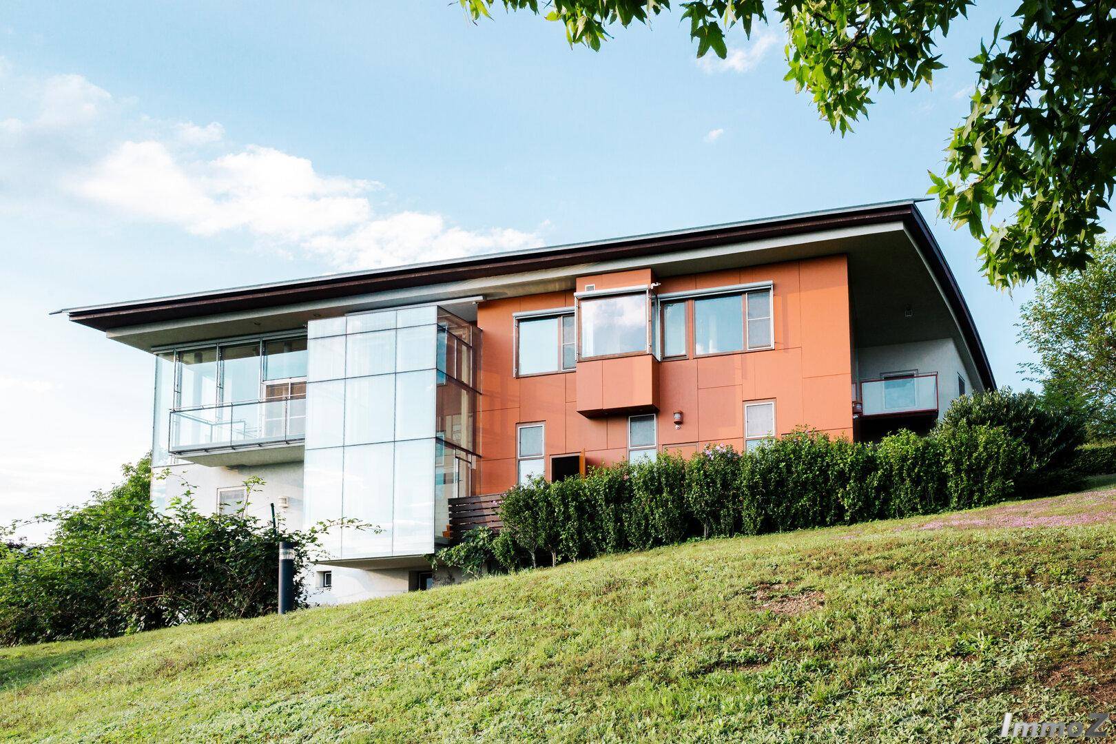 Ansicht des modernen Hauses mit orangefarbener Fassade, Glasanbau und Hanglage.