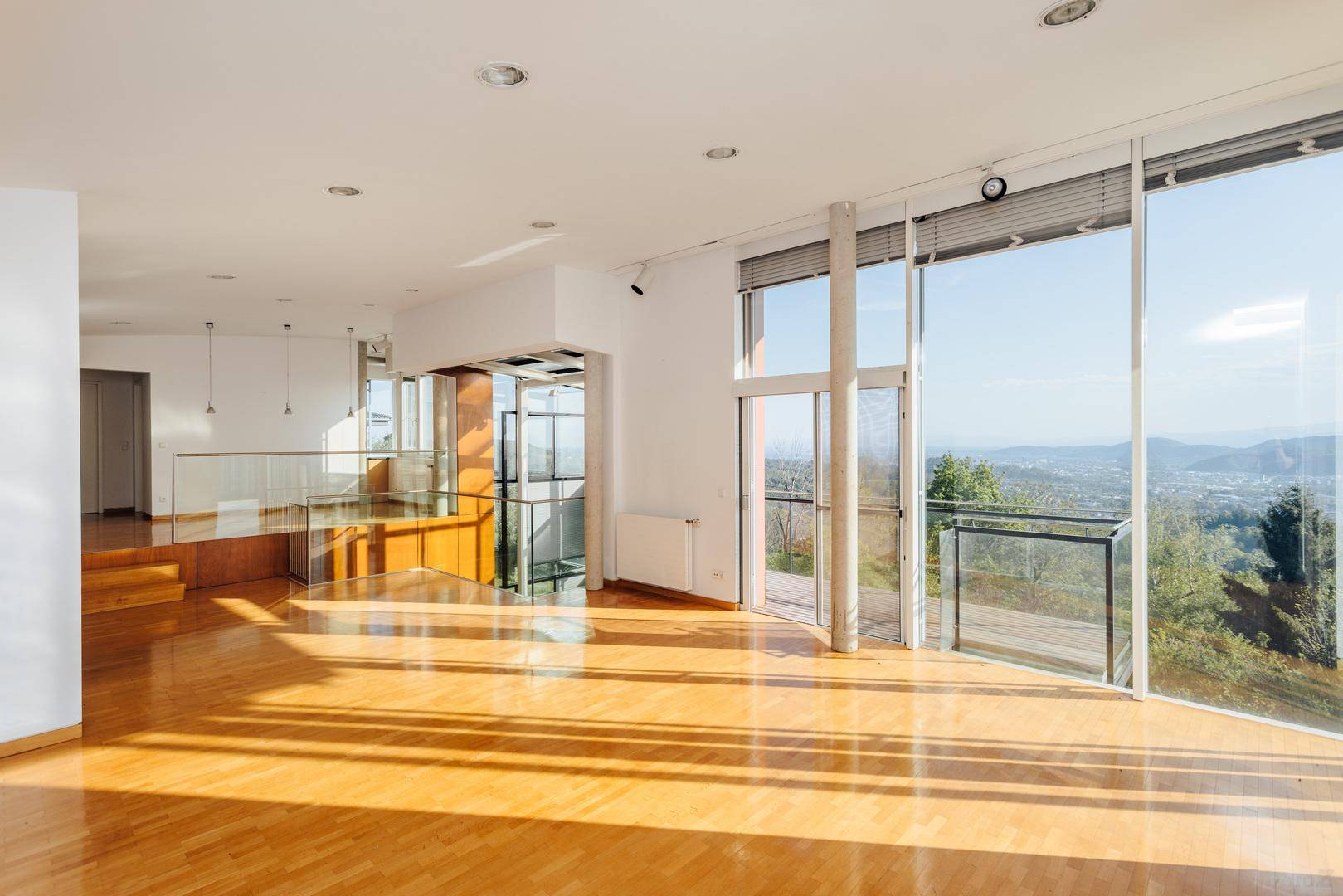 Offener Wohnbereich mit Holzboden, Glasgeländer und Zugang zu einem Balkon mit Panoramablick.