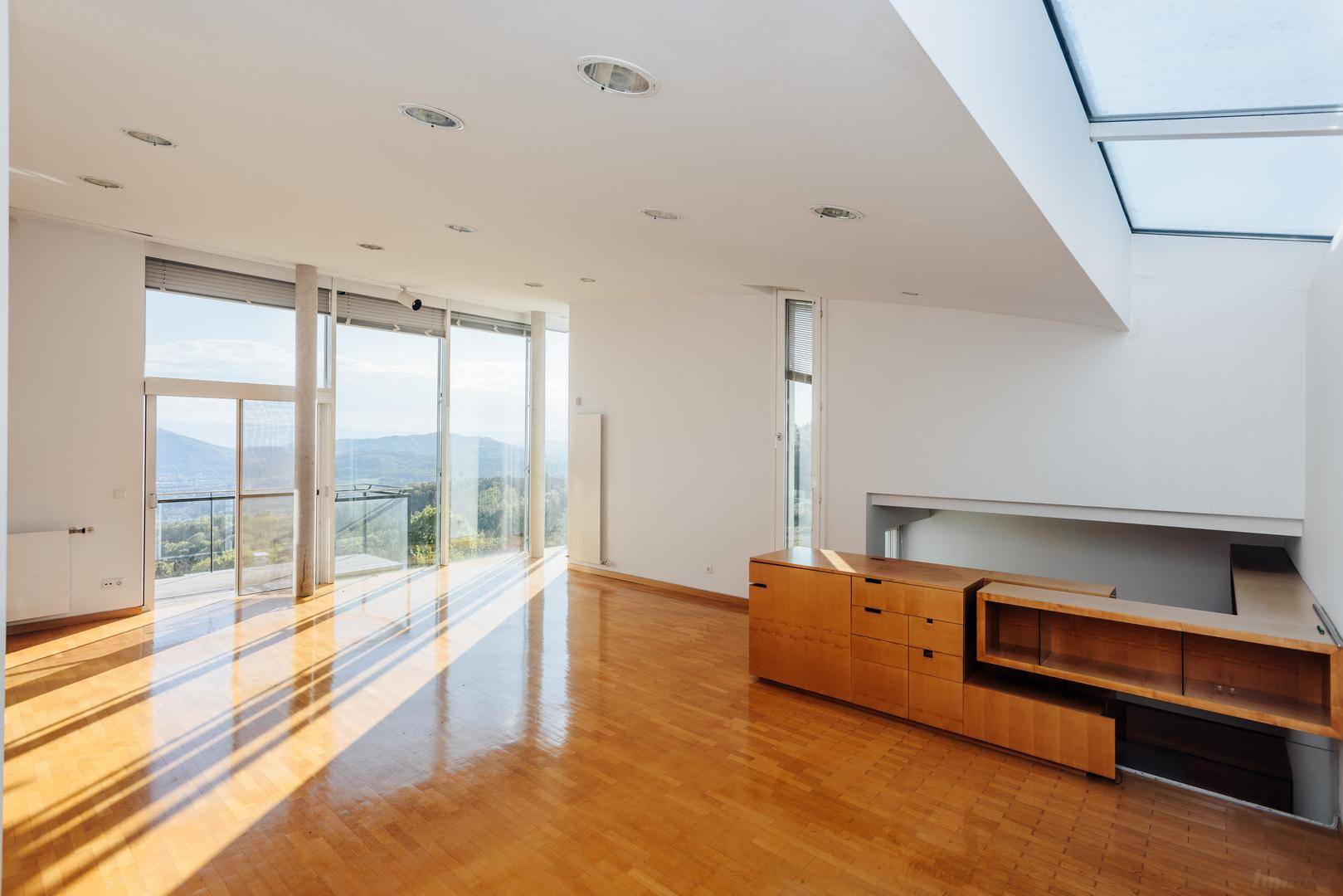 Geräumiger, lichtdurchfluteter Wohnbereich mit Holzboden, großen Fenstern und Blick auf die umliegende Landschaft.