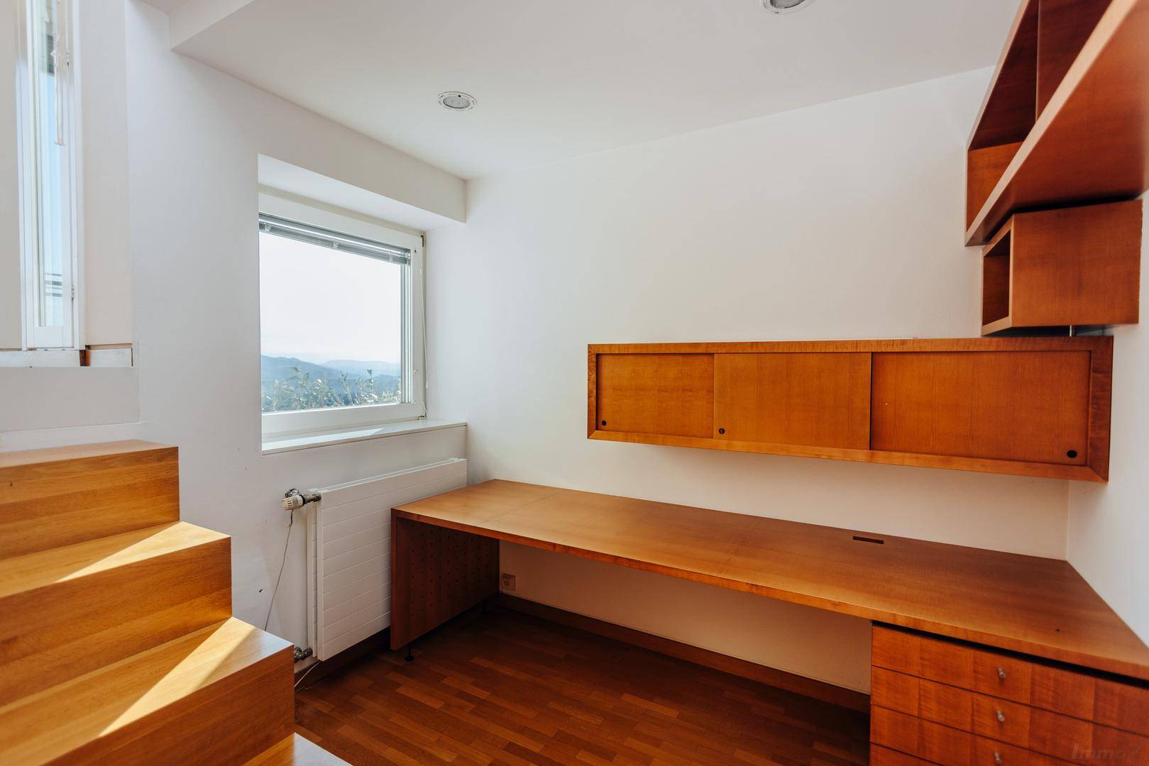 Helles Arbeitszimmer mit Holzboden, Einbauschreibtisch und Blick ins Grüne durch das Fenster.