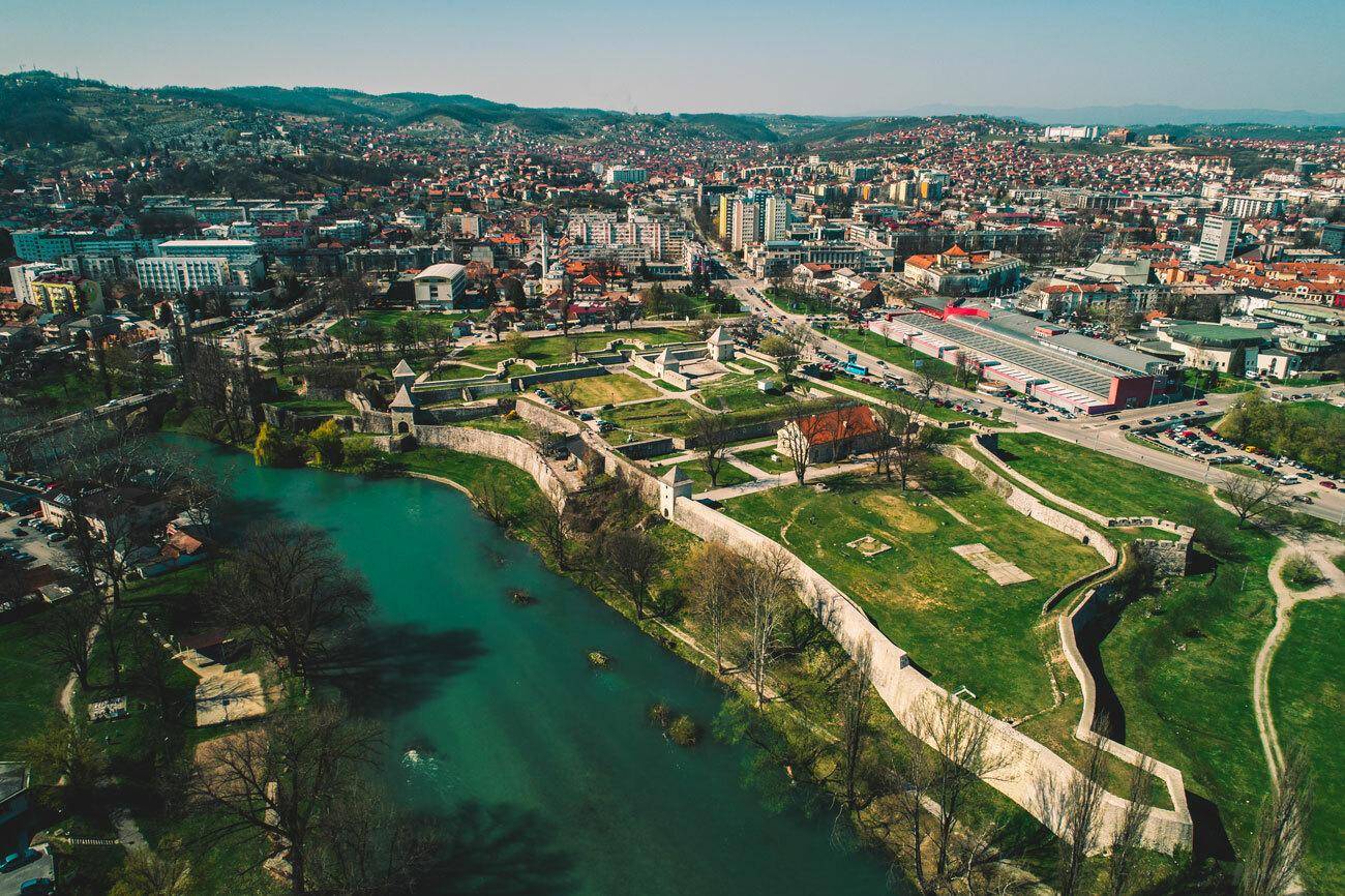 Panoramablick auf eine Stadt mit einem Fluss, historischen Festungsanlagen und umliegenden grünen Hügeln.