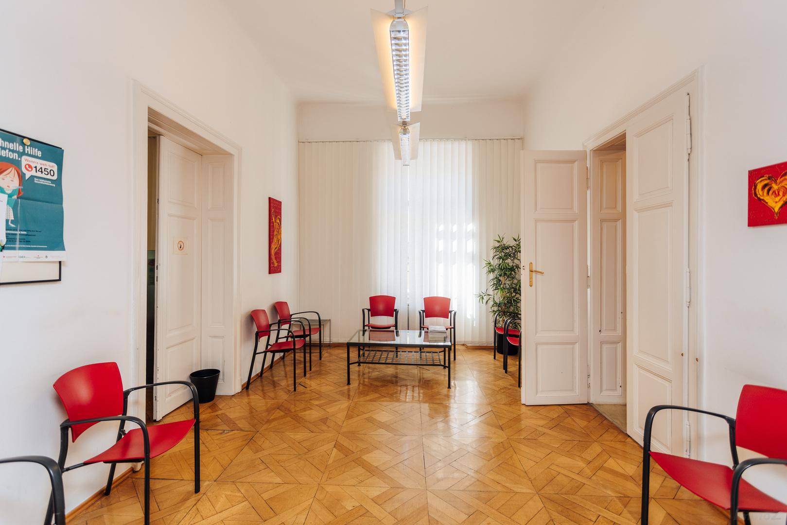 Geräumiges Wartezimmer mit Holzparkett, roten Stühlen und einem Couchtisch, hell und einladend.