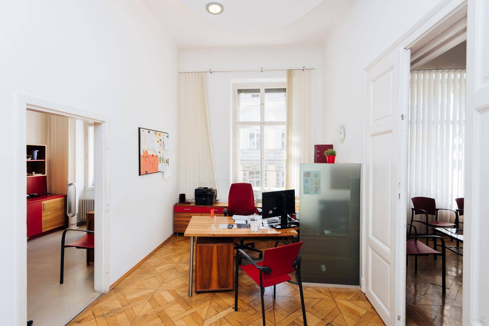Helles Büro mit Holzparkett, Schreibtisch und rotem Stuhl, mit Blick auf die Straße.