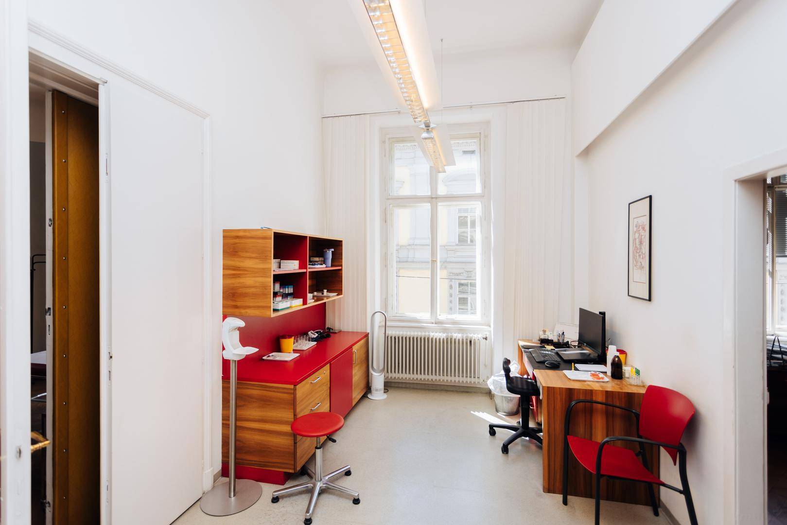 Helles Untersuchungszimmer mit Schreibtisch, roten Schränken und einem Fenster, gut ausgestattet.