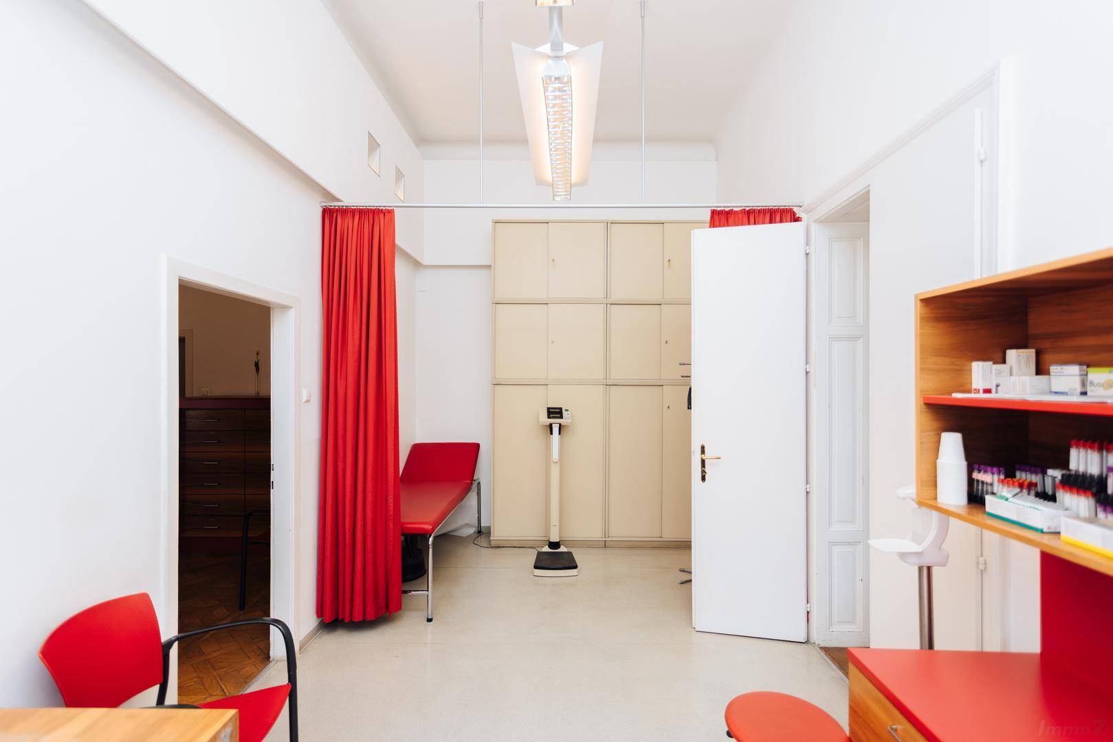 Untersuchungszimmer mit medizinischer Liege, Schränken und einer Waage, durch Vorhang abtrennbar.
