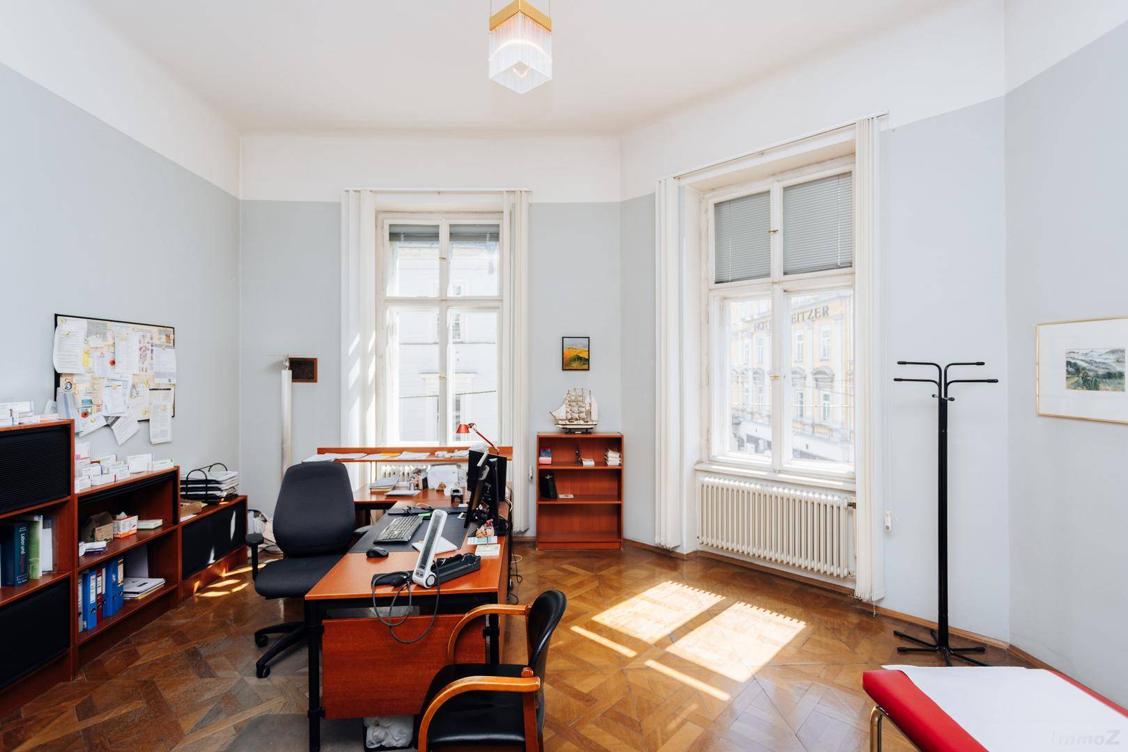 Büro oder Behandlungszimmer mit Holzparkett, Schreibtisch und medizinischer Liege, gut beleuchtet.