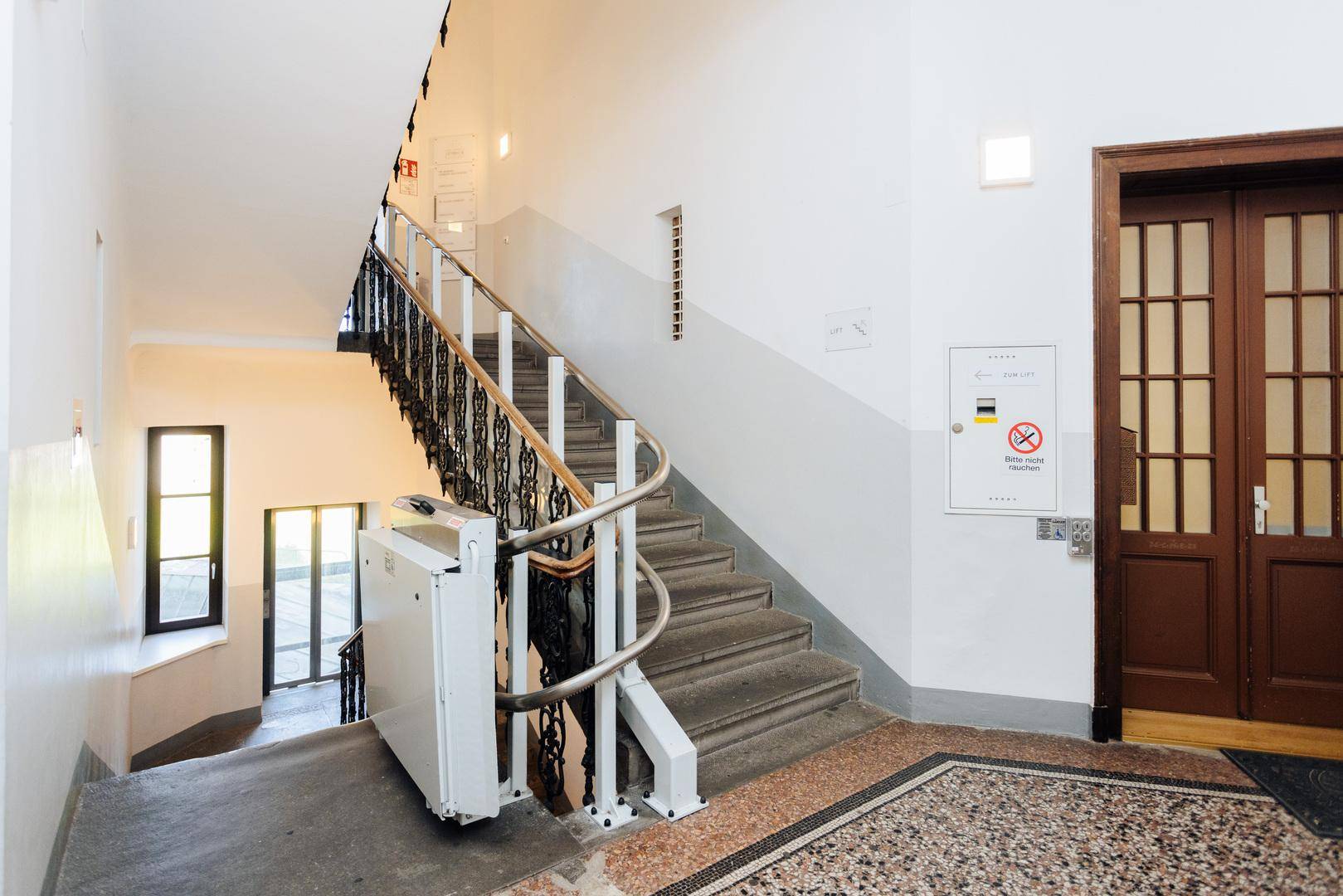 Treppenhaus mit Holzgeländer, Marmorboden und einem Treppenlift für Barrierefreiheit.
