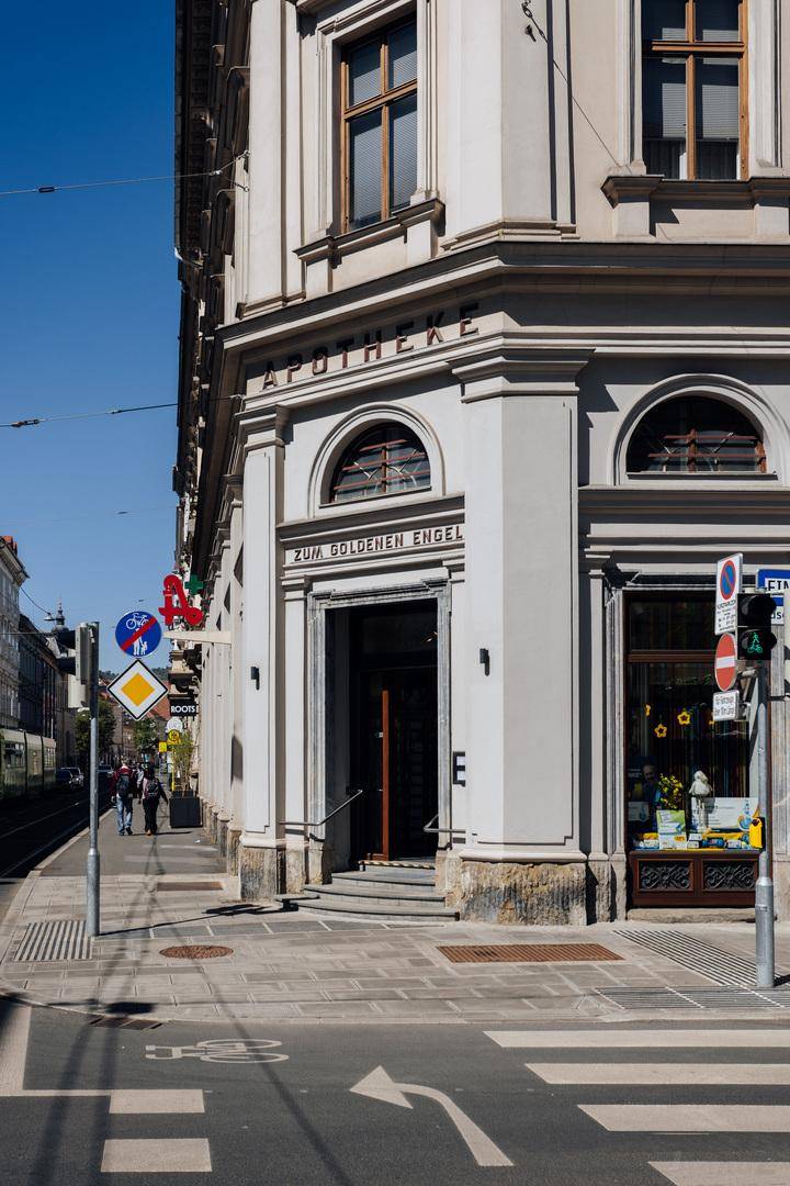 Historisches Gebäude mit Apotheke im Erdgeschoss an einer belebten Straße unter blauem Himmel.