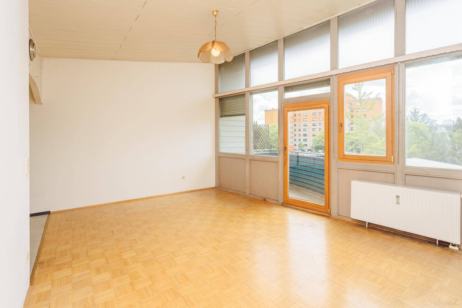 Geräumiger, lichtdurchfluteter Raum mit Parkettboden und Zugang zum Balkon, umgeben von Natur.