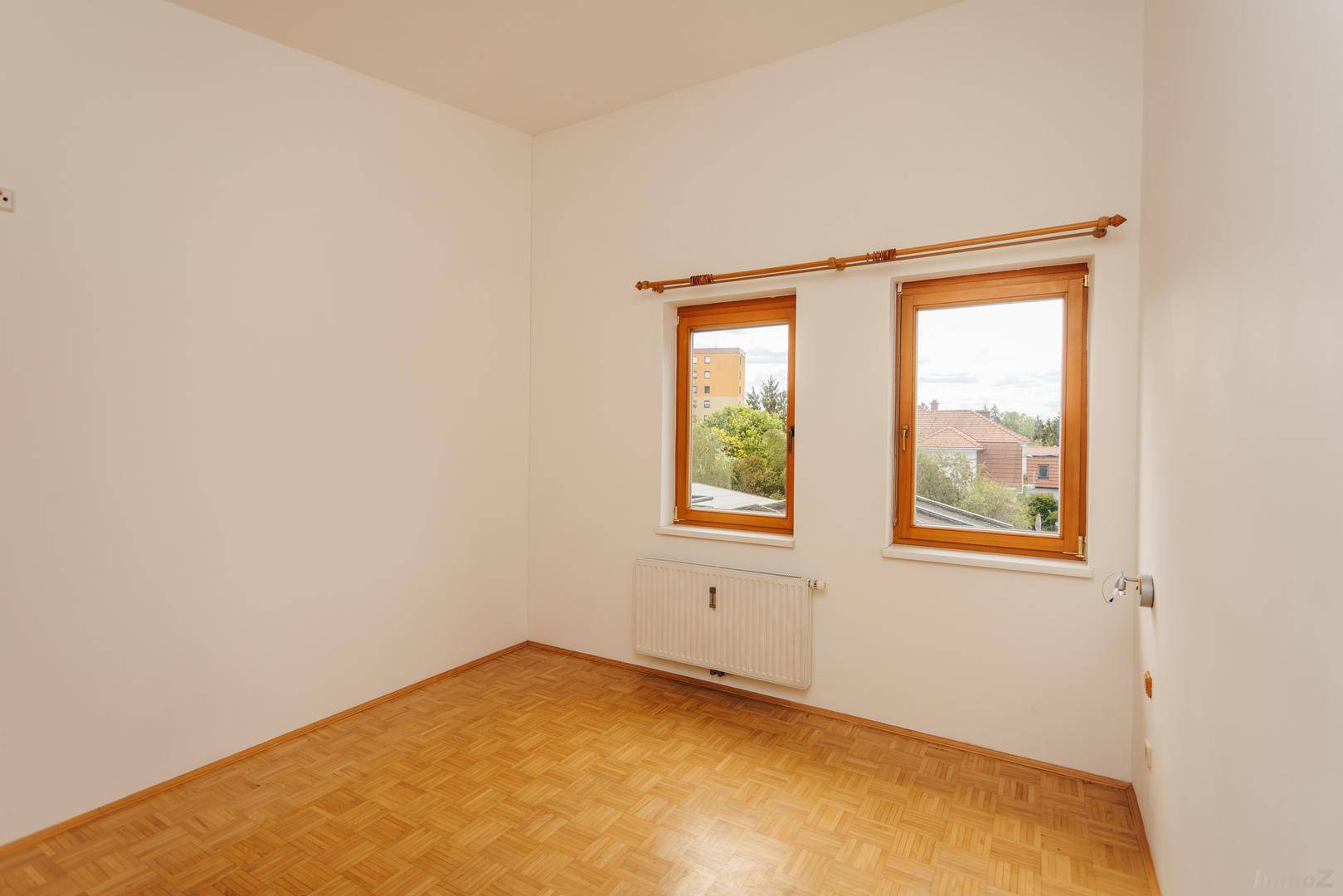 Leeres Zimmer mit Holzboden und zwei Fenstern, die viel Tageslicht hereinlassen und Ausblick bieten.
