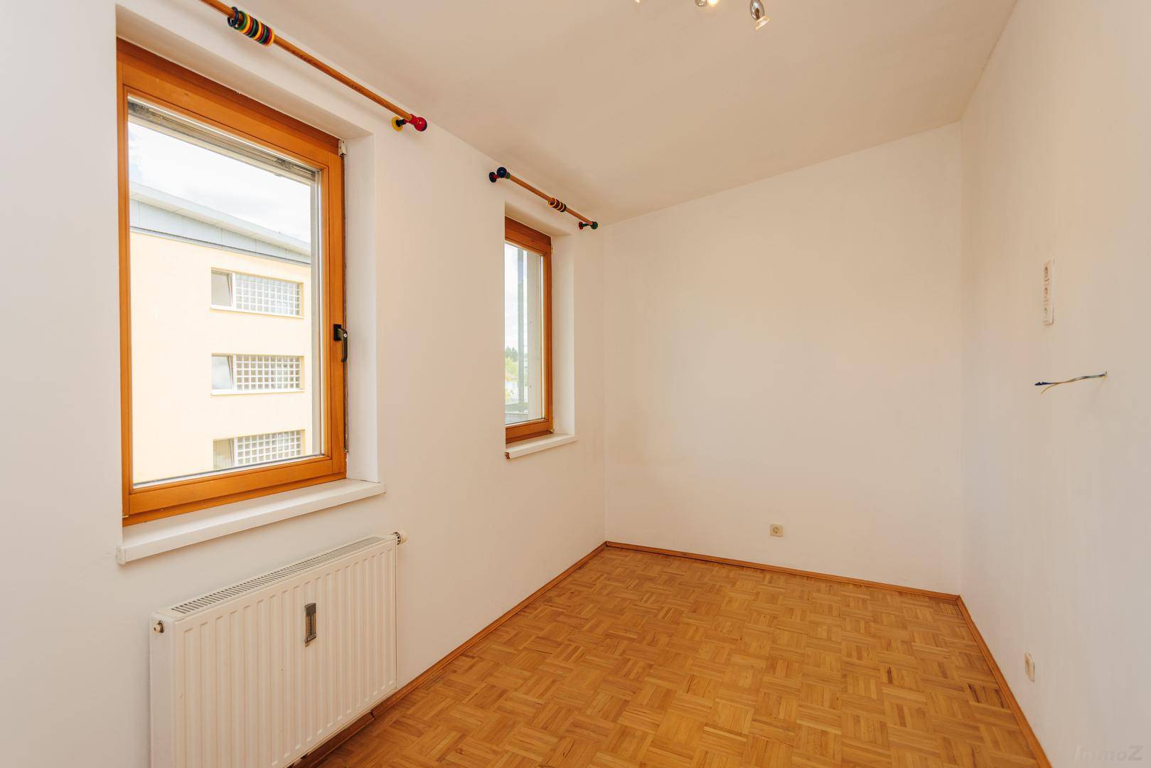 Helles Zimmer mit Parkettboden und zwei Fenstern, die einen Blick auf die umliegenden Gebäude bieten.