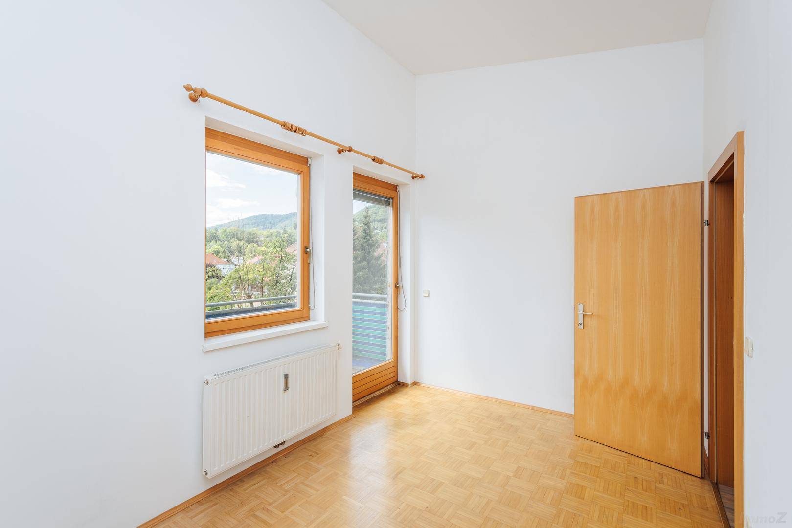Leeres Schlafzimmer mit hellem Parkettboden, großen Fenstern und Blick ins Grüne.