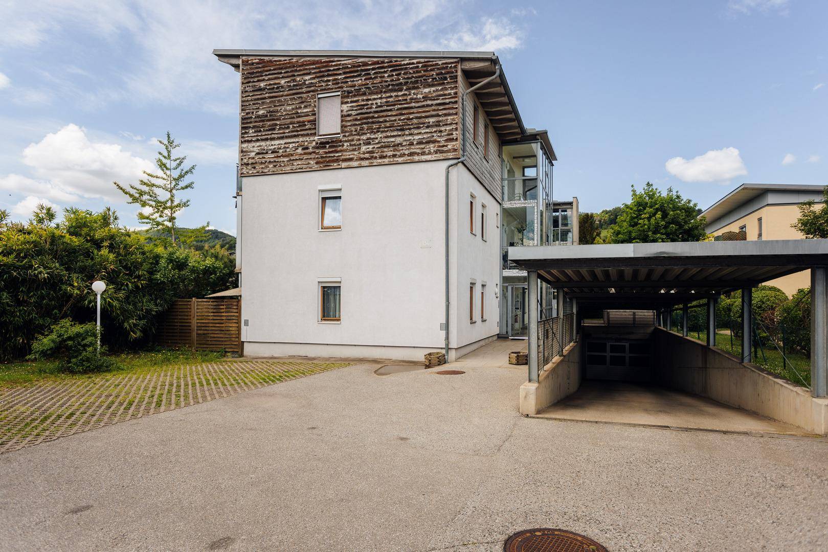 Außenansicht des Gebäudes mit Carport, Parkplätzen und umgebender Vegetation an einem sonnigen Tag.