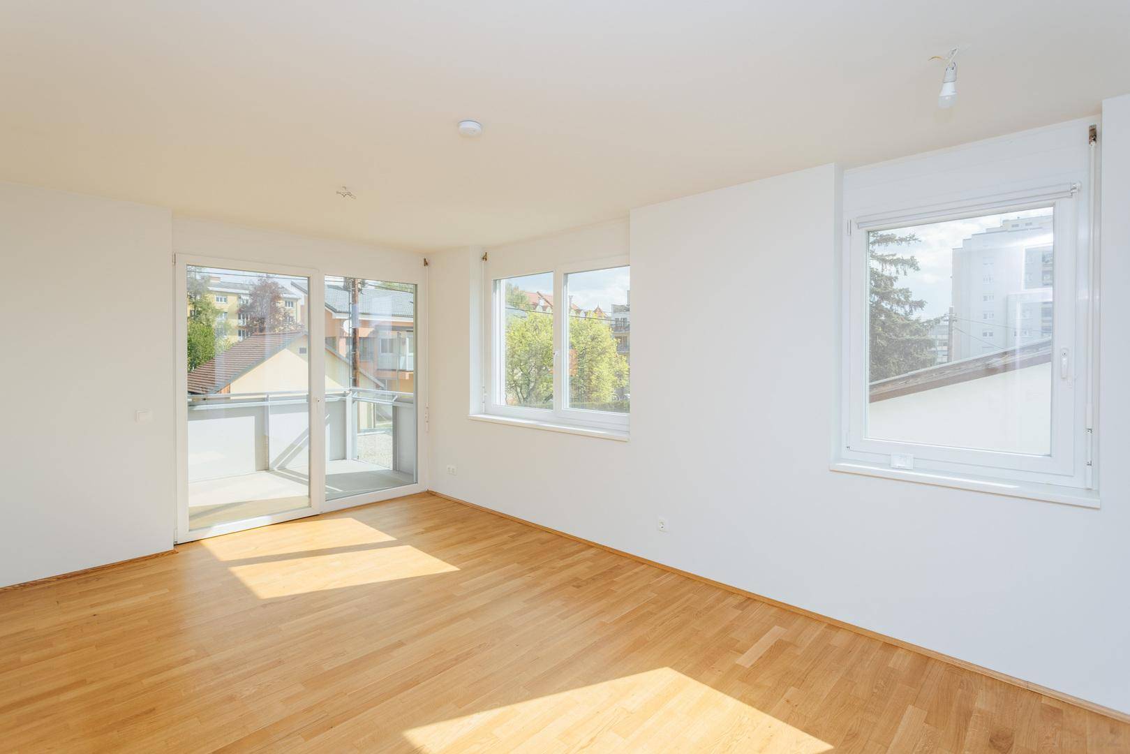 Leerer Raum mit Holzboden und großen Schiebetüren, die auf einen Balkon führen und viel Licht hereinlassen.
