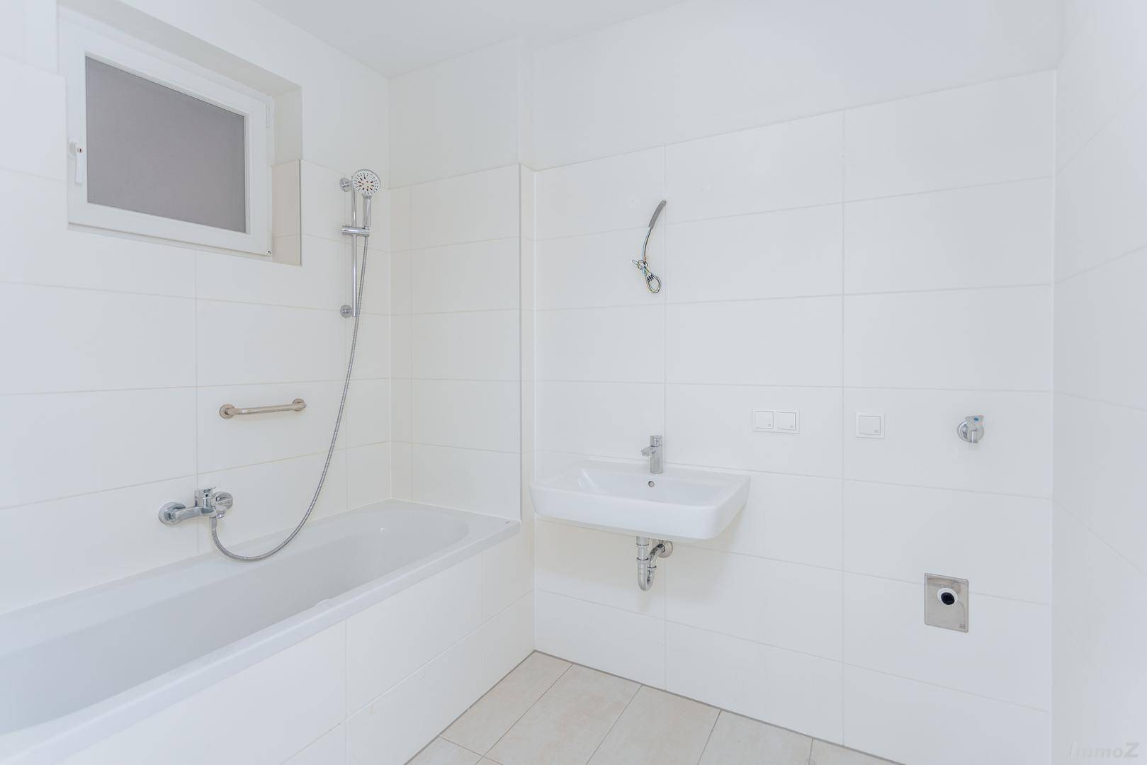 Helles Badezimmer mit weißen Fliesen, Badewanne mit Duschfunktion, Waschbecken und Fenster.