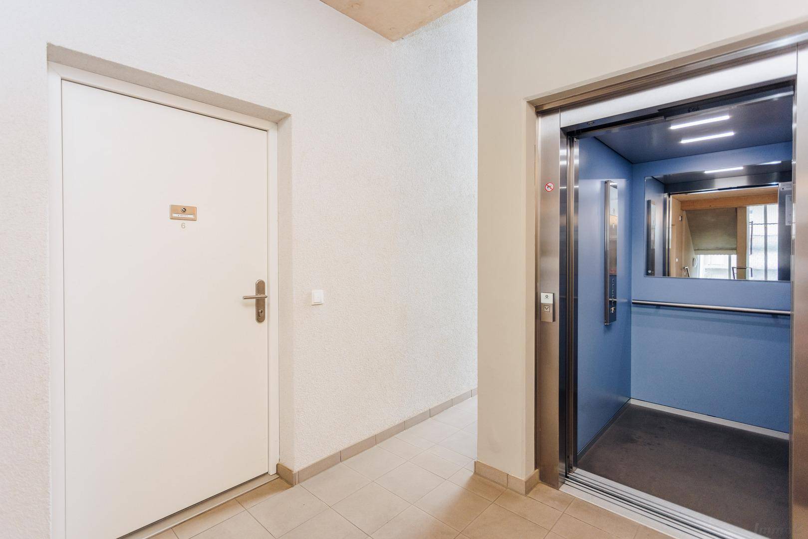Heller Flur mit Fliesenboden und Zugang zu einem modernen Aufzug mit blauer Kabine.