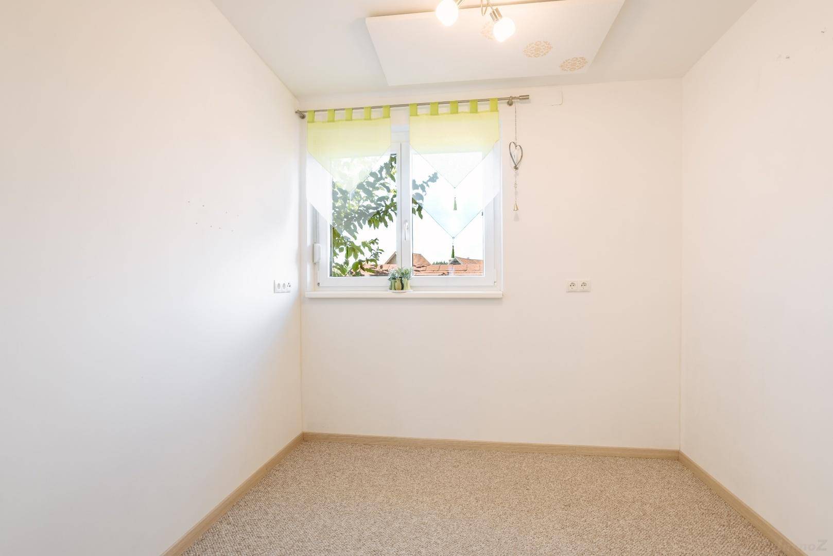 Helles, leeres Zimmer mit Teppichboden und einem Fenster, das viel Tageslicht hereinlässt.