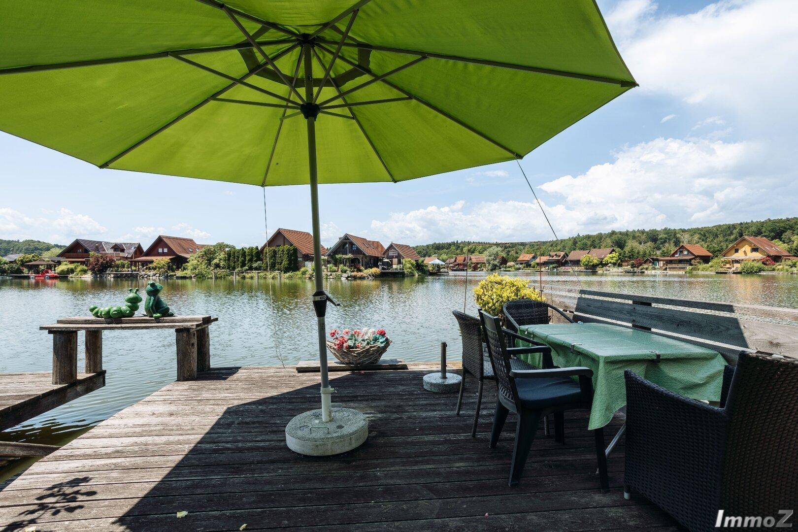 Gemütliche Holzterrasse direkt am See mit Esstisch, Stühlen und großem Sonnenschirm.
