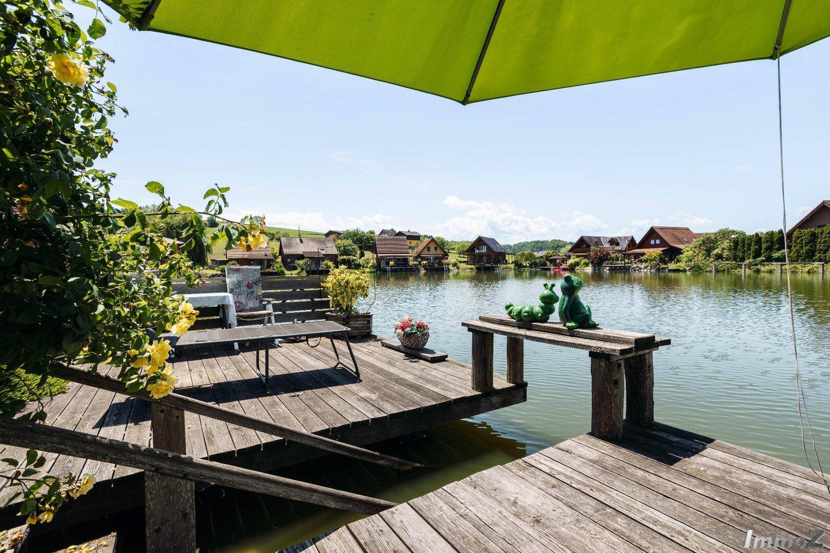 Weitläufige Holzterrasse am Seeufer mit Sitzgelegenheiten und Blick auf die umliegenden Häuser.