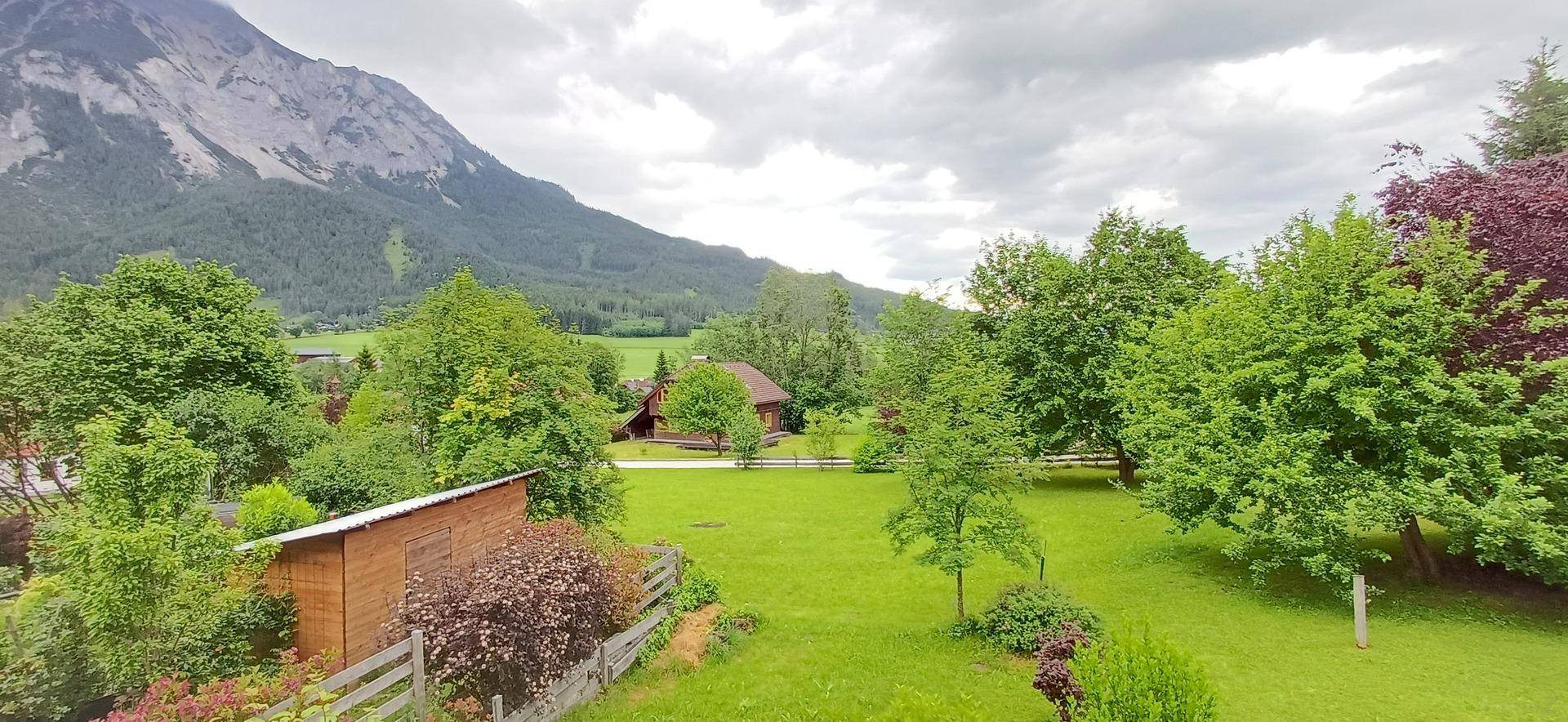 Weitläufiger Blick über grüne Wiesen, Bäume und ein kleines Haus mit majestätischen Bergen im Hintergrund.