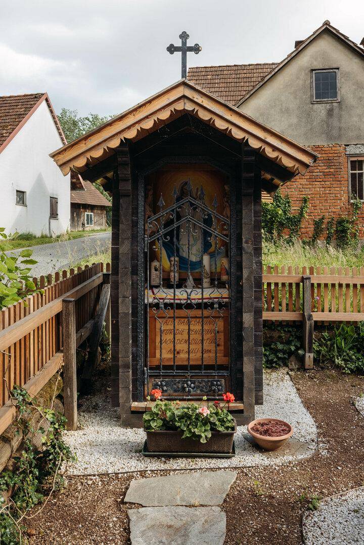 Kleiner religiöser Schrein mit Kreuz und Blumen in einem ländlichen Vorgarten.