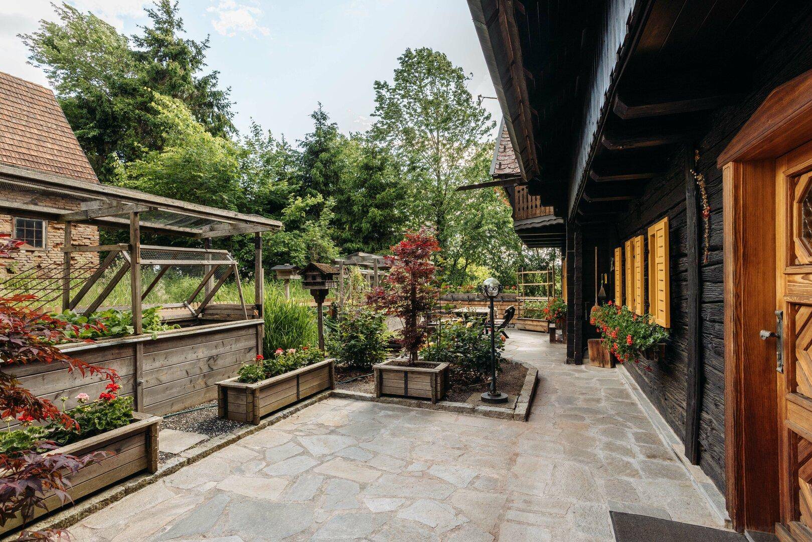 Langer gepflasterter Terrassenbereich entlang des Hauses mit Holzpflanzkästen und grüner Umgebung.