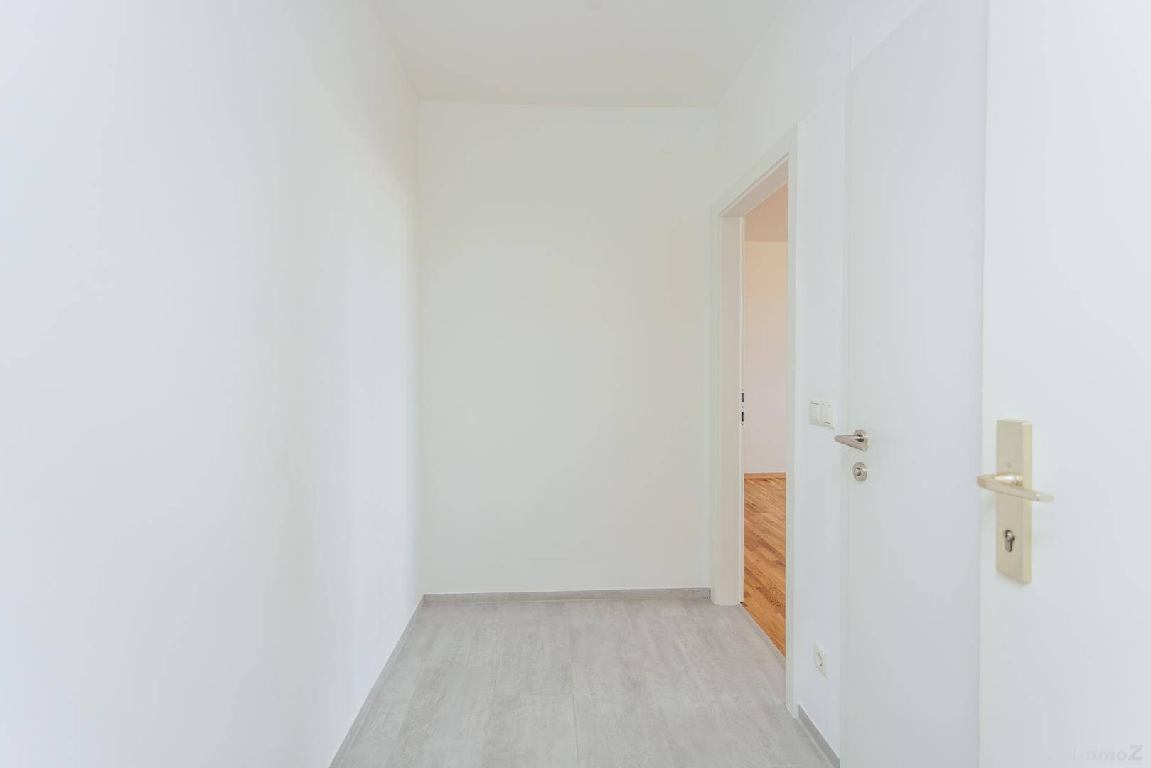 Leerer, heller Flur mit hellgrauen Fliesen und weiß gestrichenen Wänden, führt zu einem Raum mit Holzboden.