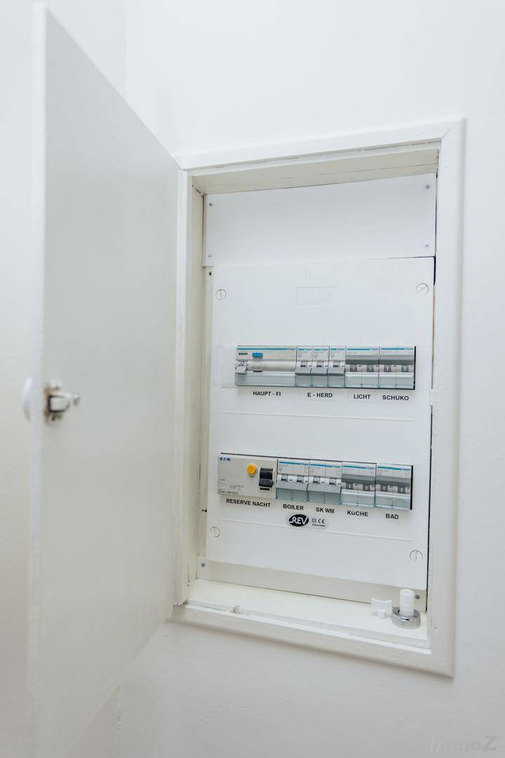 Offener Sicherungskasten mit verschiedenen Schaltern und Beschriftungen an einer weißen Wand.
