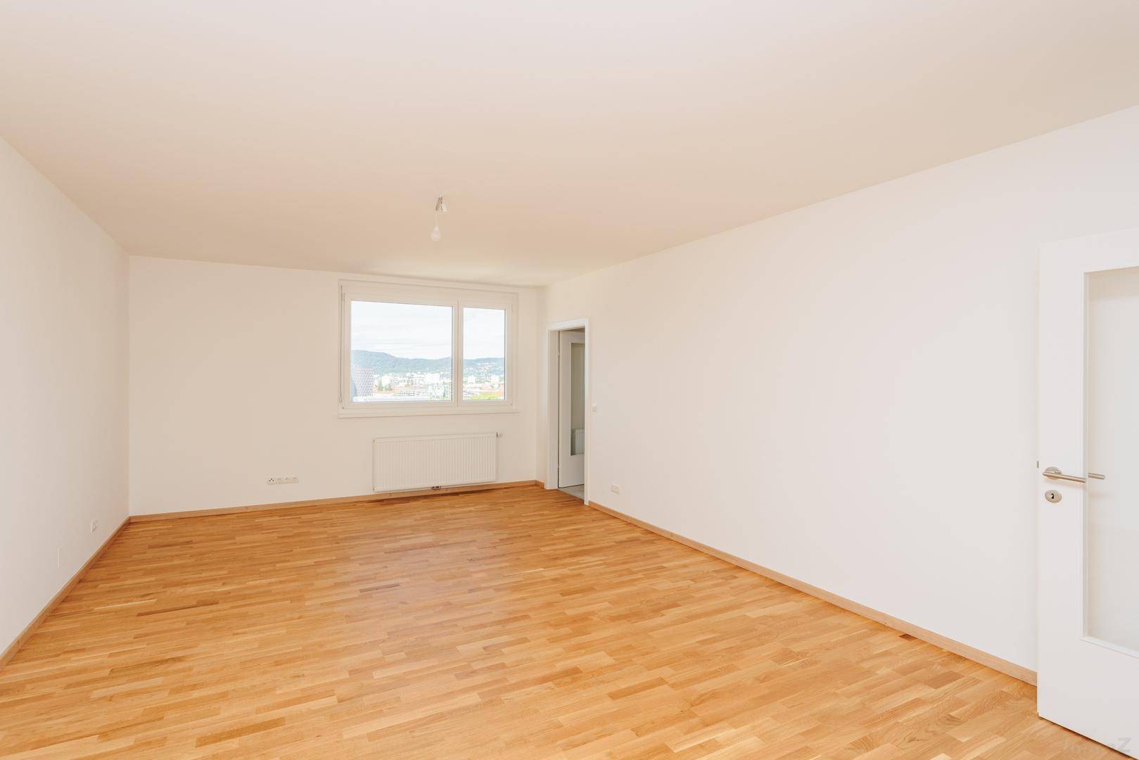 Geräumiger, leerer Raum mit Holzboden, weißen Wänden und weitem Blick auf die Stadt.