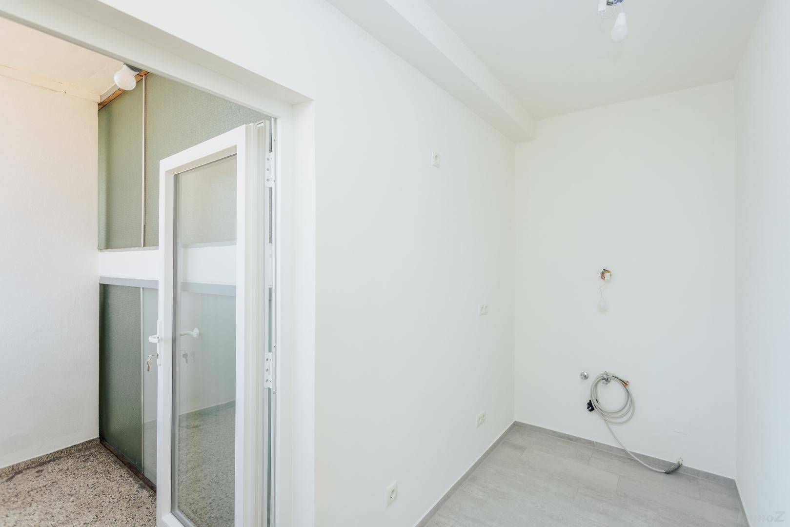 Leerer Küchenbereich mit Fliesenboden, weißen Wänden und Anschlüssen für eine Einbauküche, mit Balkonzugang.