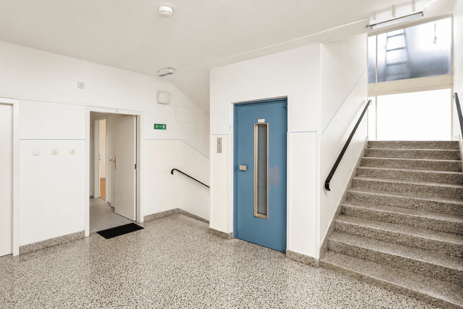 Heller Hausflur mit Terrazzoboden, Treppenaufgang und einem blauen Aufzug im Gebäude.