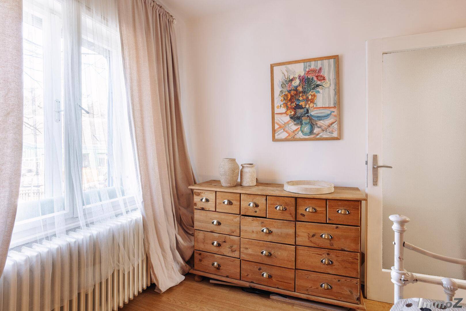 Schlafzimmer mit Holzkommode, großem Fenster und hellem Parkettboden.
