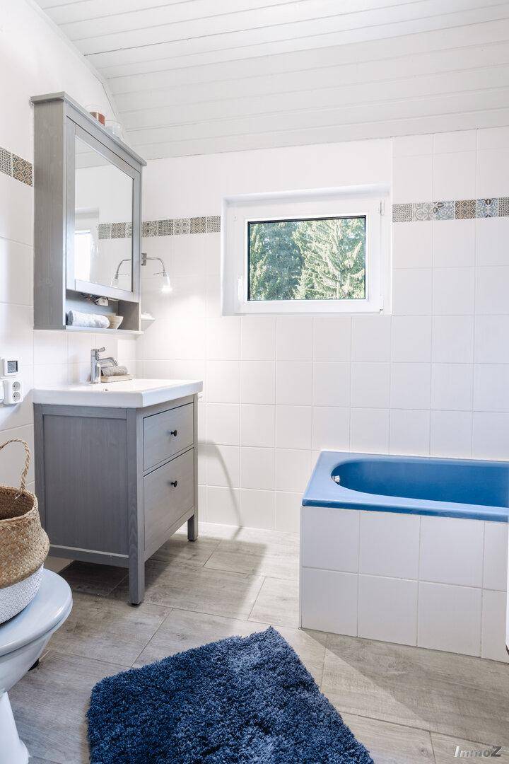 Modernes Badezimmer mit Waschtisch, Spiegel und blauer Badewanne unter einem Fenster.