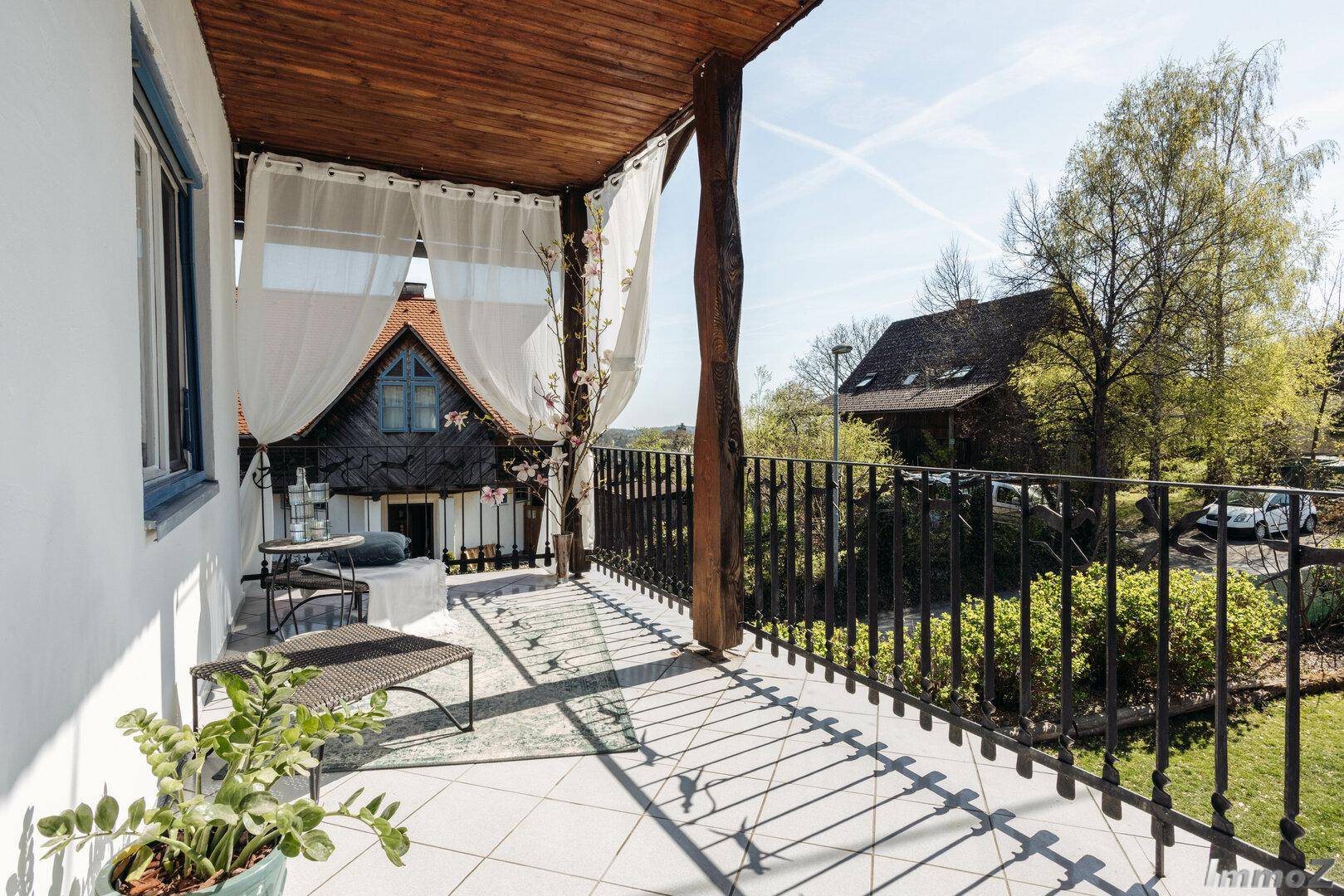 Überdachter Balkon mit schmiedeeisernem Geländer, Fliesenboden und Blick ins Grüne.