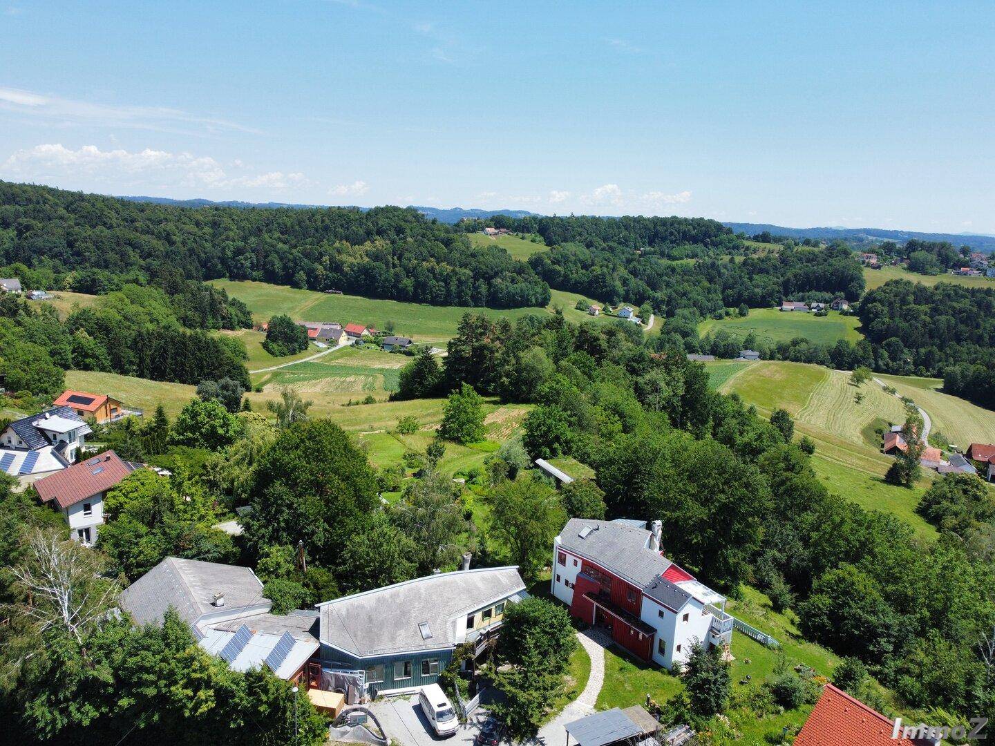 Luftaufnahme einer Siedlung mit mehreren Häusern, eingebettet in eine grüne, hügelige und bewaldete Landschaft.