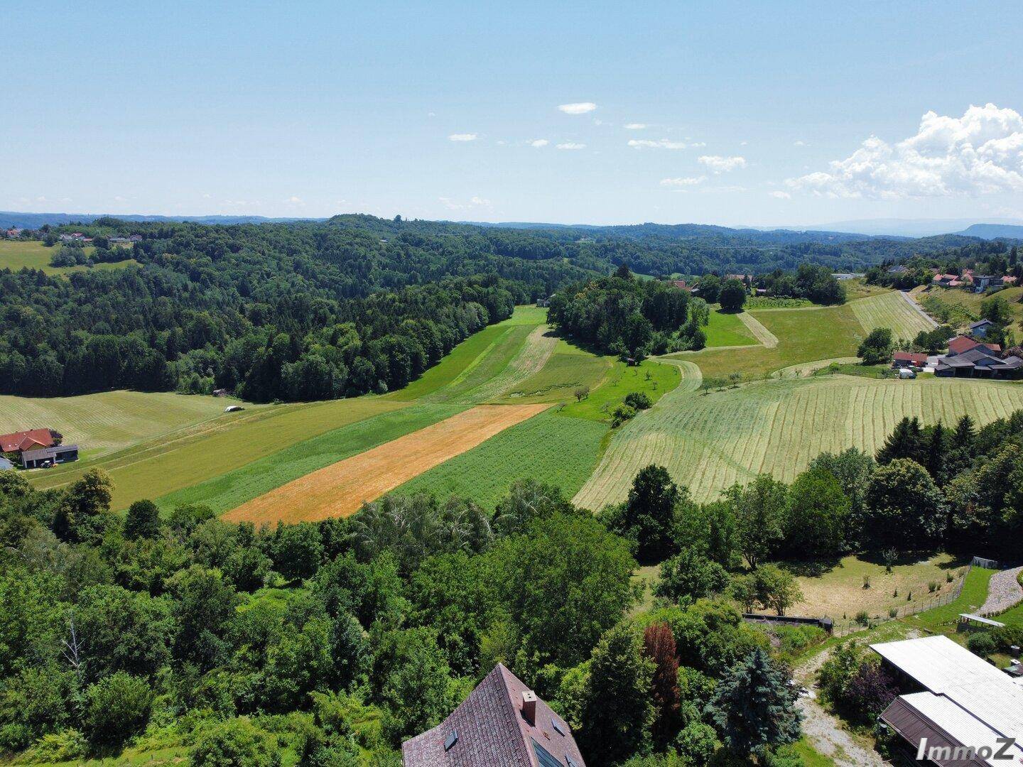 Luftaufnahme einer weitläufigen ländlichen Landschaft mit Feldern, Wäldern und vereinzelten Häusern unter blauem Himmel.