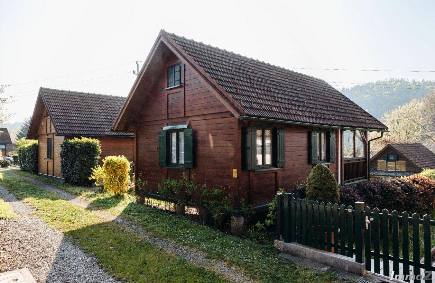 Holzhaus mit grünen Fensterläden und einem kleinen Vorgarten, umzäunt von einem Holzzaun.