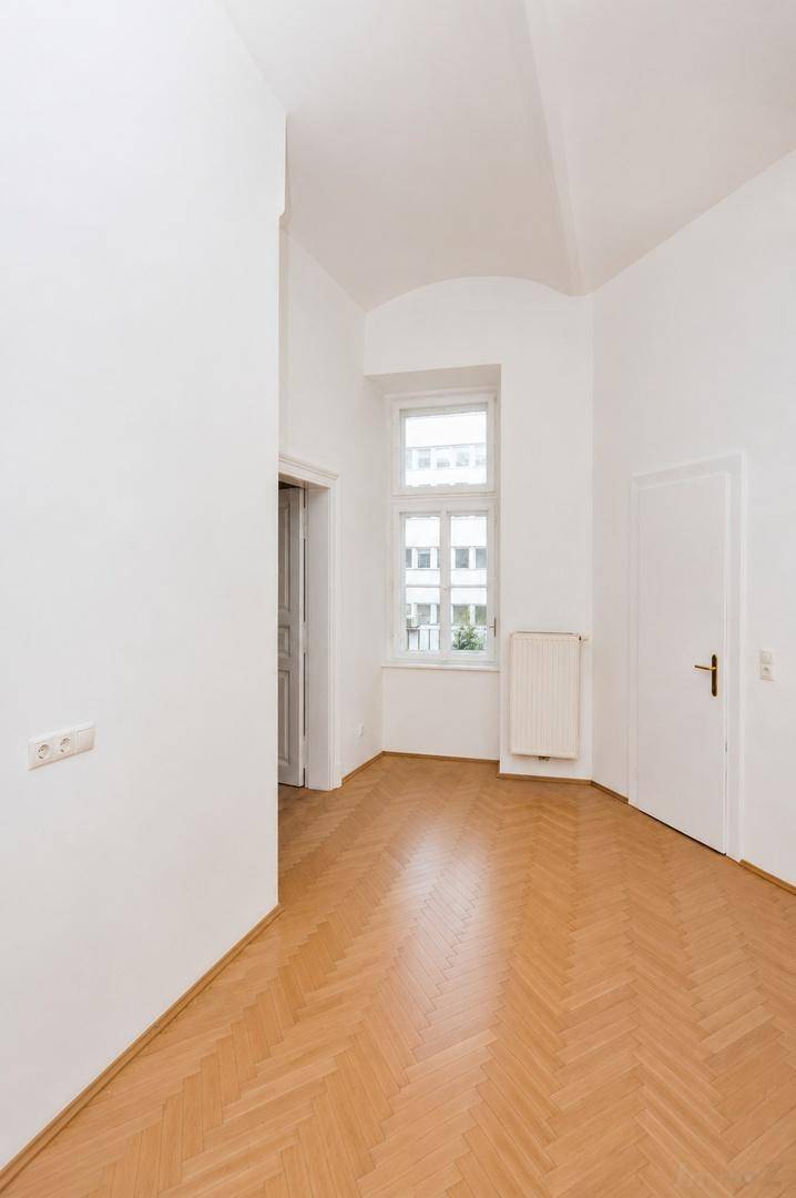 Leerer Raum mit weiß gestrichenen Wänden, Fischgrätparkett und einem Fenster, das Tageslicht hereinlässt.