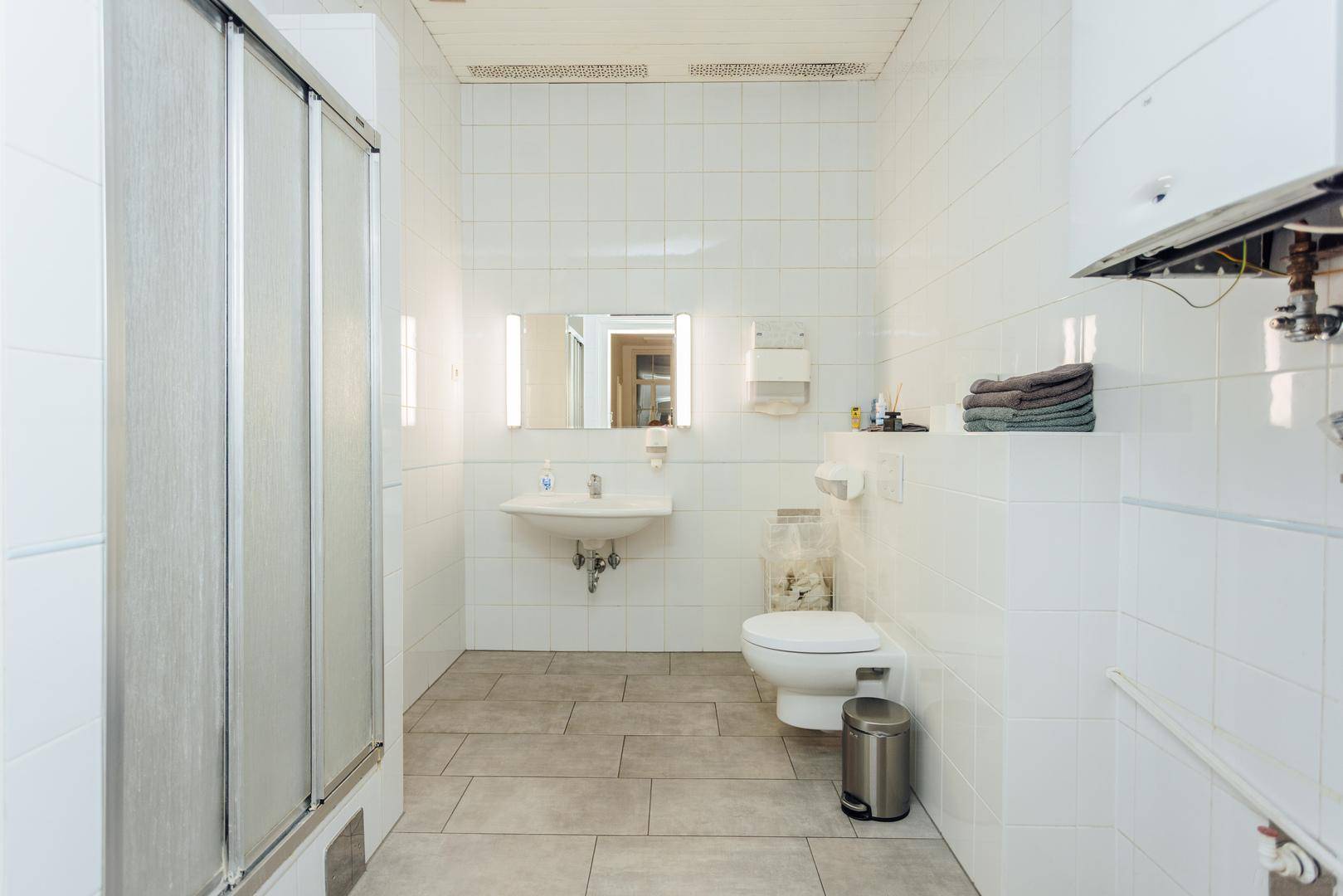 Helles Badezimmer mit weißen Fliesen, Duschkabine, WC und einem Wandspiegel mit Beleuchtung.