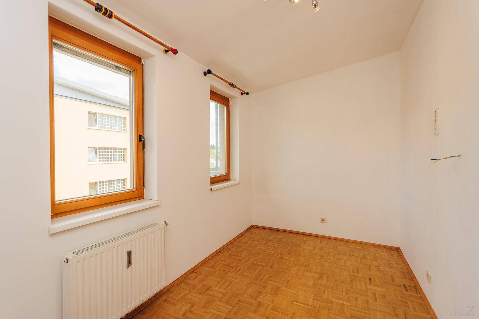 Leeres Zimmer mit Parkettboden und zwei Fenstern, die viel Tageslicht hereinlassen.