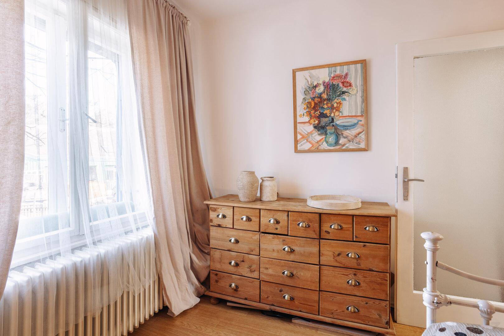 Schlafzimmer mit Holzboden, Fenster und einer antiken Holzkommode als Blickfang.