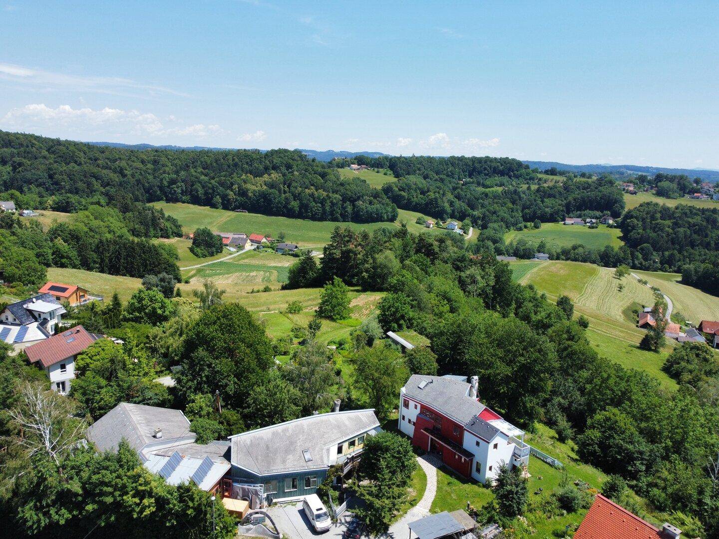 Luftaufnahme einer kleinen Siedlung inmitten einer grünen, hügeligen Landschaft mit Wäldern und Feldern.