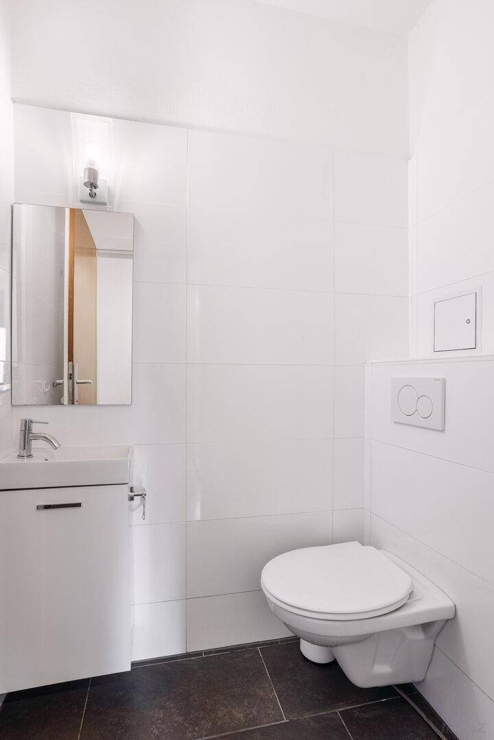 Modernes Gäste-WC mit weißen Fliesen, wandhängendem WC und kleinem Waschbecken.