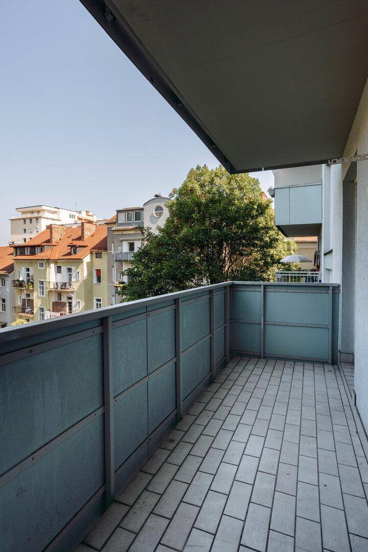 Geräumiger Balkon mit Fliesenboden und Blick auf Bäume und Gebäude.