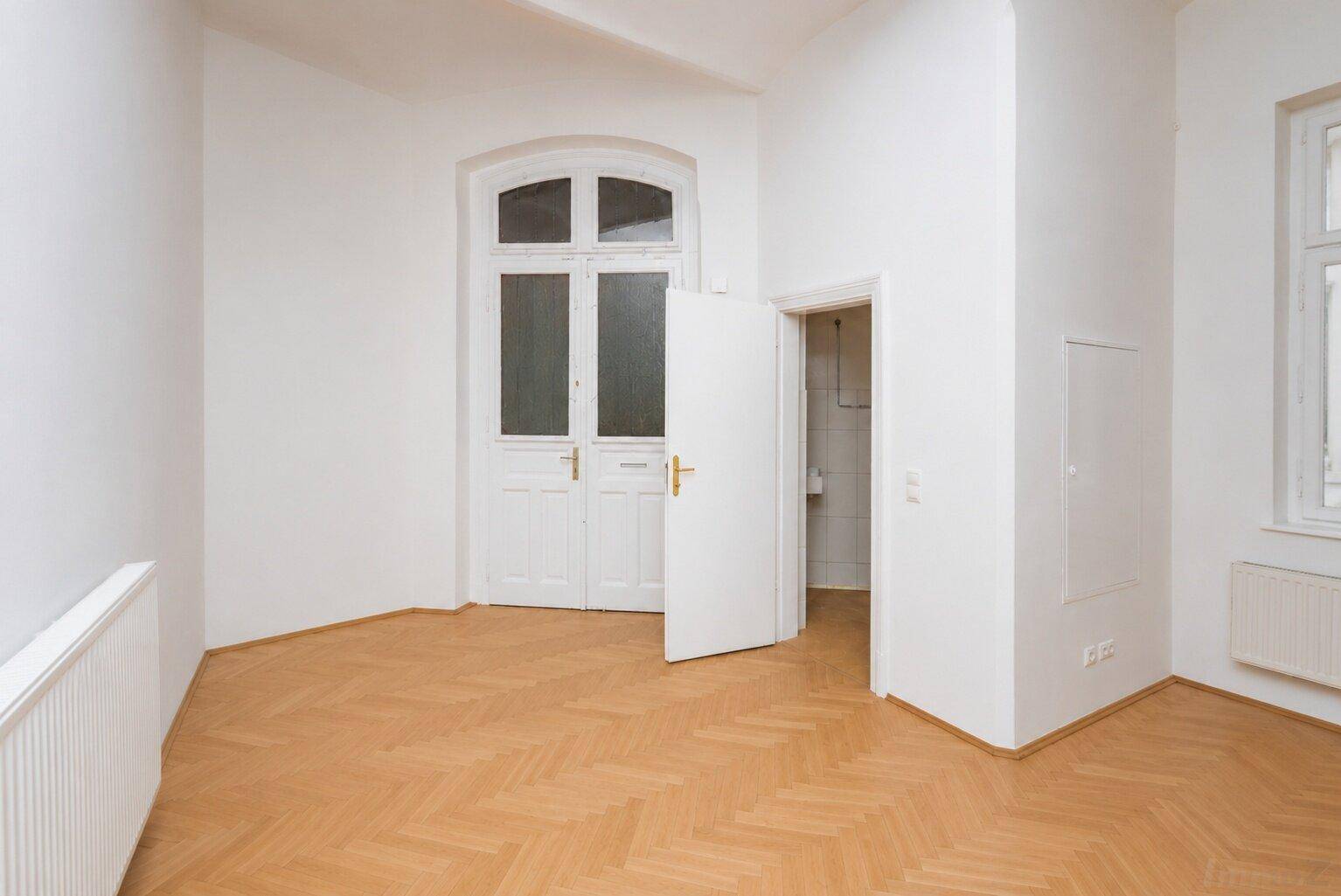 Heller Raum mit Fischgrätparkett, hohen Türen und Zugang zu einem weiteren Zimmer.