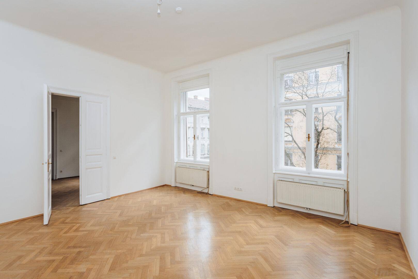 Geräumiger Raum mit Fischgrätparkett, zwei hohen Fenstern und weiß gestrichenen Wänden.