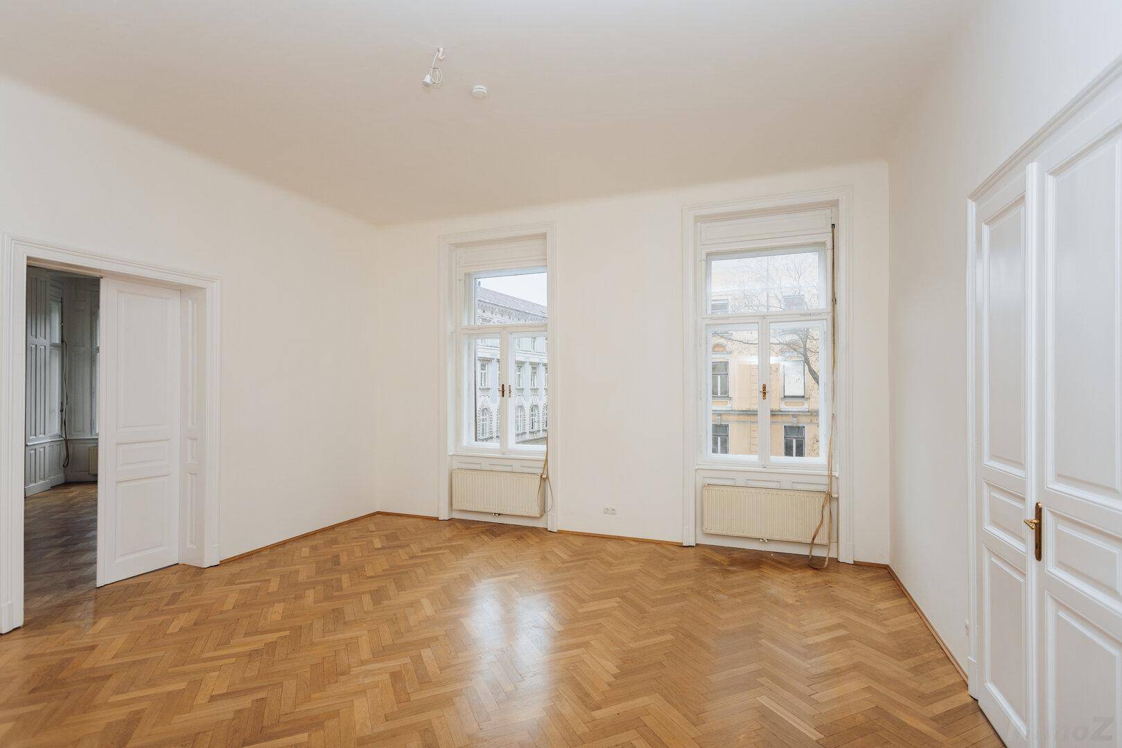 Heller Raum mit Fischgrätparkett, zwei Fenstern und einer Doppeltür zu einem weiteren Zimmer.