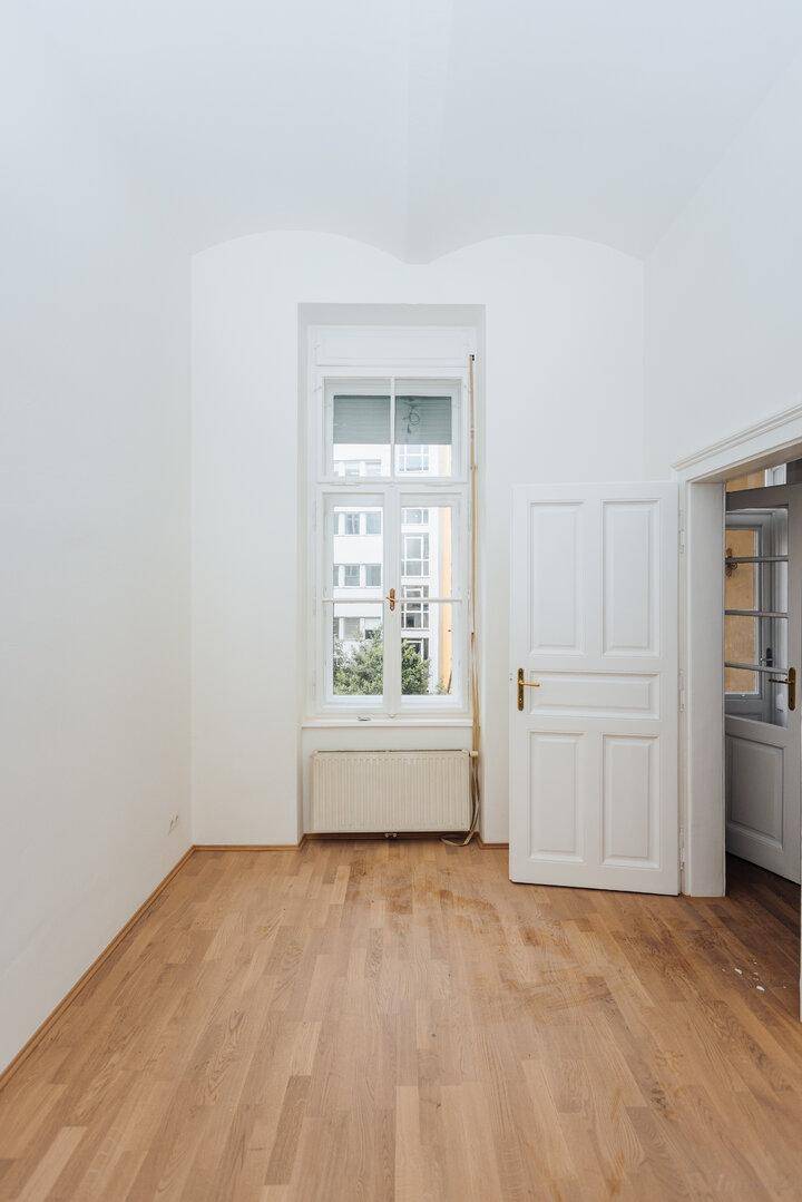 Heller Raum mit Fischgrätparkett, einem hohen Fenster und einer geöffneten Tür.