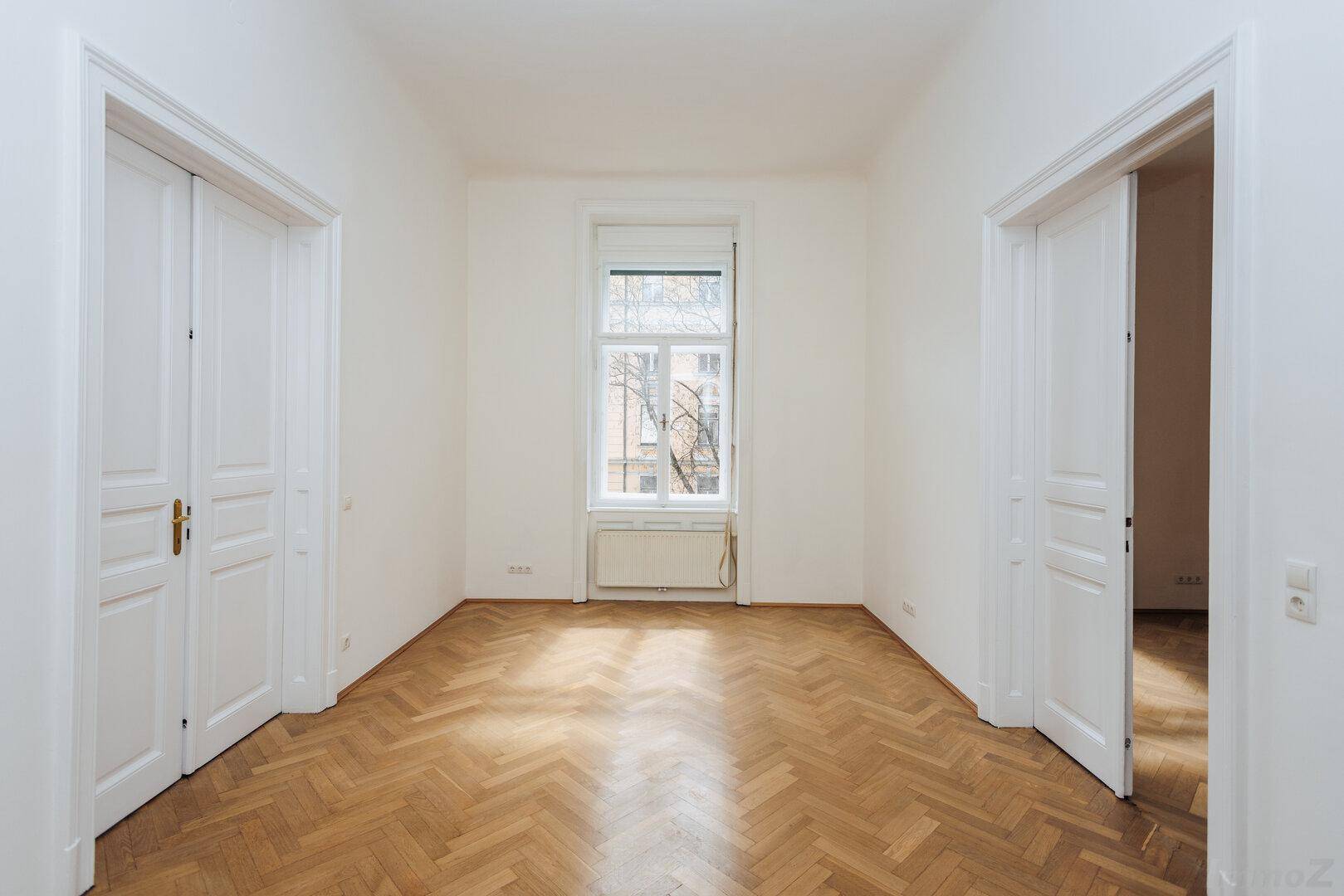 Langer Raum mit Fischgrätparkett, einem Fenster und zwei gegenüberliegenden Türen.
